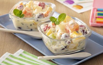 Fotografia de dois recipientes quadrados pequenos de vidro com salada de frutas e creme de maracujá com ninho em cada um, misturado. Por cima deles tem algumas folhas de hortelã, ao lado tem uma colher clara, também sobre um apoio retangular azul.