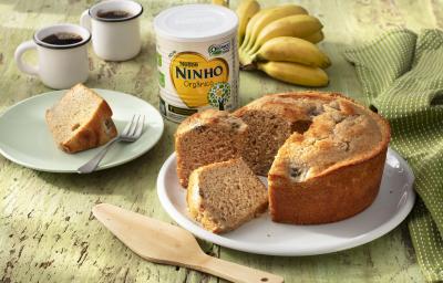 Fotografia em tons de verde de uma bancada verde com um prato branco redondo, sobre ele um bolo de banana. Ao lado um prato com uma fatia de bolo e um garfo, duas xícaras de café, um paninho verde com um cacho de banana e uma lata de NINHO Orgânico