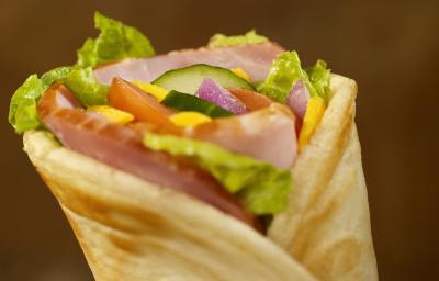 Fotografia de um lanche fechado com pão sírio, alface, tomate, pepino, cebola, molho e peito de peru, e um fundo escuro.