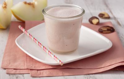 Fotografia em tons de branco e rosa de uma bancada branca com um paninho rosa e um prato quadrado branco, sobre ele um copo de vidro com o Smoothie e um canudo listrado rosa. Ao fundo duas fatias de melão e castanhas-do-pará.