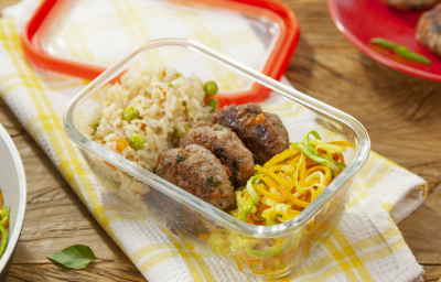 Fotografia em tons de amarelo em uma mesa de madeira com um pano de prato branco com listras amarelas, um potinho de marmita de vidro com o arroz integral, o mini hambúrguer e o macarrão de legumes