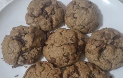 Fotografia de sete cookies grandes com gotas de chocolate sobre um prato grande e raso, de cor branco.