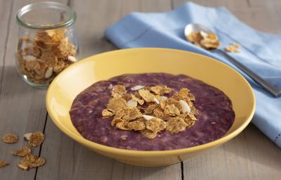 Fotografia em tons de roxo em uma bancada de madeira escura com um pano azul claro ao lado, um prato redondo fundo amarelo com o creme de açaí dentro dele e decorado com cereais. Ao fundo, um potinho de vidro com cereais.