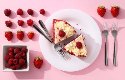 Fotografia vista de cima de um prato raso grande branco de vidro com duas fatias de torta red velvet, com dois garfos juntos ao lado. Ao redor do prato tem mais dois garfos e muitas frutas vermelhas.