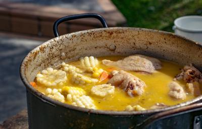 Fotografia de uma panela funda e grande com um ensopado de frango com espigas de milho e cenoura. A panela está apoiada sob alguma mesa ao ar livre.