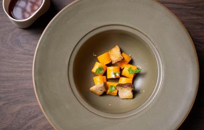 Fotografia vista de cima de um prato escuro com o meio fundo. Dentro do prato tem uma receita de nhoque de batata doce laranja, dashi de missô e cogumelos tostados. O prato está sobre uma bancada de madeira escura.