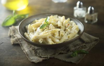 Fotografia em tons de cinza de uma bancada de madeira, ao centro um paninho cinza amarronzado com um prato redondo sobre ele com o macarrão pene com molho branco.