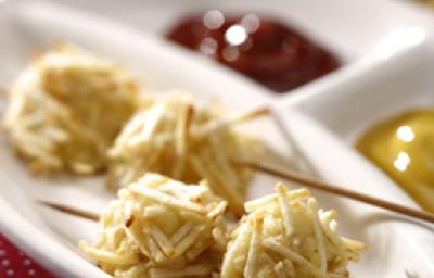 Fotografia em tons de vermelho em uma mesa com uma toalha vermelha com bolinhas brancas, um recipiente branco com o espetinho de cachorro quente empanado com batata palha, e ketchup e mostarda ao lado.