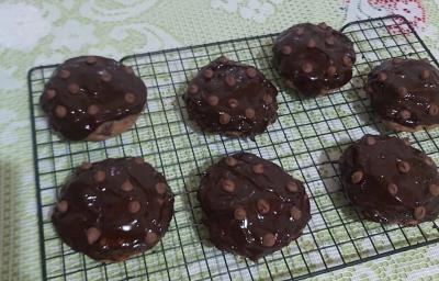 Foto da receita de Cookies low carb de chocolate. Observa-se uma grade preta com 7 cookies dispostos em cima, com calda de chocolate e chocolate em gotas na cobertura.