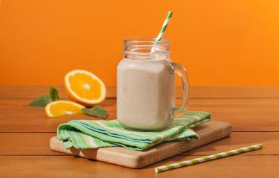 Fotografia em tons de laranja em uma bancada de madeira, um fundo laranja. Ao centro, uma tábua de madeira, um paninho verde, um canudo de papel verde e em cima um copo grande de vidro com o shake de banana dentro. Ao fundo, uma laranja aberta e folhas.