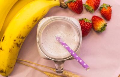 Fotografia em tons de rosa, de uma bancada rosa vista de cima, ao centro um copo com uma bebida e um canudo, em volta bananas e morangos.