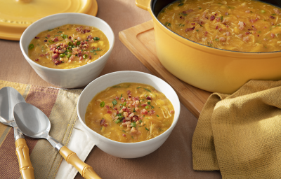 Foto em tons de amarelo da receita de caldo de kenga servida em dois bowls brancos de porcelana com duas colheres prateadas ao lado. Ao lado uma panela com o caldo e um paninho, ambos amarelos, sobre uma tábua de madeira. Ao fundo, a tampa da panela