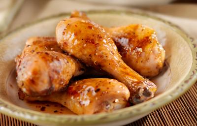 Fotografia de cinco coxas de frango refogadas com um brilho do caldo dentro de um recipiente fundo de vidro de cor clara, que está sobre um apoio de mesa com decoração de madeira.