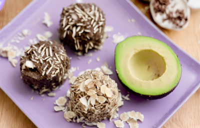 Fotografia em tons de roxo com um prato retangular roxo ao centro. Em cima do prato existe 3 brigadeiros de cor marrom e ao lado existe metade de um abacate. Todos os brigadeiros estão polvilhados com coco ralado