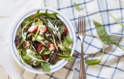 Fotografia vista de cima de um recipiente fundo branco com uma listra azul claro na borda, com uma salada de folhas, com molho de creme de leite e tomates cereja. O recipiente está sobre duas toalhas de mesa, nas cores branco e azul, e ao lado, um garfo.