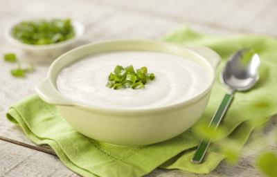 Fotografia em tons de verde em uma bancada de madeira com um guardanapo de pano verde ao centro com uma tigelinha branca e o creme de couve-flor dentro enfeitado com cebolinhas verdes. Ao lado, uma colher e ao fundo um potinho com cebolinhas verdes.