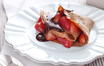 fotografia em tons de branco, marrom e vermelho de uma bancada cinza vista de frente, contém um prato branco redondo com um Crepe de Chocolate com Morangos e ao lado uma colher para servir.