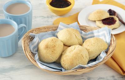 Foto da receita de Pão de Leite em Pó. Observa-se uma cestinha com um guardanapo azul e branco com 5 pãezinhos dentro. Ao lado esquerdo, duas canecas com café com leite. Do direito, um pratinho com um pão cortado ao meio recheado de geleia.