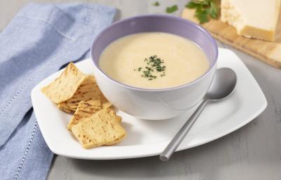 Fotografia em tons de cinza, branco e azul de uma bancada cinza com um prato branco retangular, sobre ele um recipiente redondo branco com o creme de queijo e torradinhas. Ao lado um paninho azul. Ao fundo uma tábua com um pedaço de queijo e salsinha.