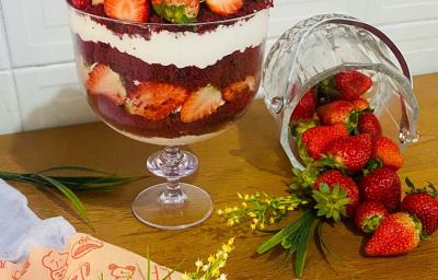 Foto em tons de vermelho da receita de red velvet na taça servida em uma graned porção em uma taça de vidro alta em cima de uma mesa de madeira com um pote de vidro com morangos ao lado