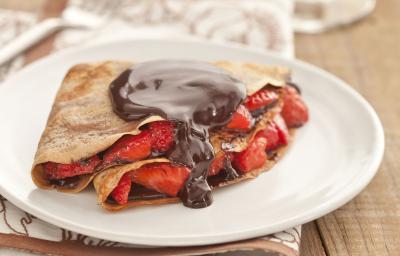 Fotografia em tons de marrom em uma bancada de madeira com um pano branco com listras marrom, um prato branco raso com o crepe recheado com morangos e chocolate.