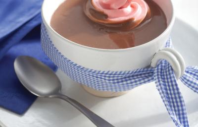 Fotografia em tons de azul em uma bancada de madeira branca com um pano azul ao lado, um prato branco raso pequeno, um xícara branca e o chocolate quente com marshmallow dentro.