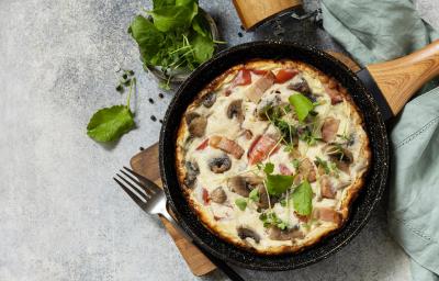 Fotografia em tons de azul claro e branco om uma frigideira ao centro. Dentro da frigideira existe um omelete feito com carne, cogumelos e algumas folhas