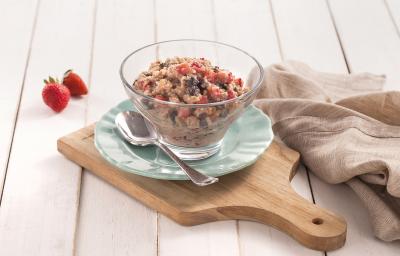 Fotografia em tons de azul em uma bancada de madeira clara, uma tábua de madeira, um paninho bege, um prato azul redondo pequeno, uma taça de vidro com o doce de quinoa com frutas secas, morango e iogurte dentro dele.