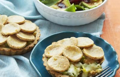 Fotografia em tons de azul em uma mesa de madeira laranja com um pano azul e um prato redondo fundo branco com salada e um prato fundo azul com a torta de massa de grão-de-bico com vegetais