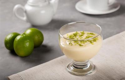 Fotografia em tons de branco com uma taça de sobremesa de vidro ao centro dentro da taça existe uma receita de mousse de limão coberta com raspas de limão.