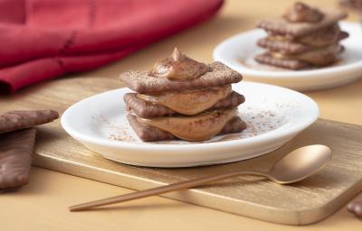 Foto de uma mesa marrom clara. Bem ao centro está uma tábua de madeira marrom clara com um prato branco contendo a receita de Tiramisù de ChocoBiscuits polvilhado com chocolate em pó. Há uma colher dourada, biscoitos espalhados e um guardanapo de pano.