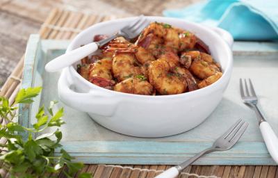 Fotografia em tons de azul em uma bancada de madeira clara, uma bandeja de madeira azul, um pano azul e um recipiente redondo branco com os camarões fritos dentro dele e em cima da bandeja. Ao lado, dois garfos.