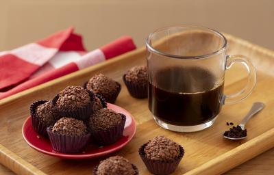 Fotografia em tons de marrom em uma bandeja de madeira de cor marrom. No centro, uma xícara de café preto e ao lado, alguns brigadeiros de Nescafé e uma colher contendo pó de Nescafé. Ao fundo, um pano vermelho.