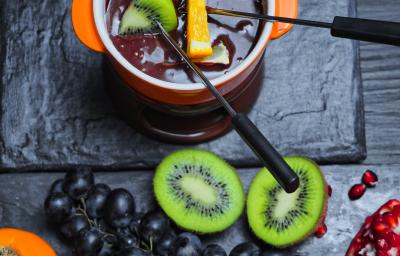 Fotografia de um Fondue de chocolate meio amargo dentro de um recipiente de vidro na cor laranja, com frutas sobre o creme. Ao lado, algumas frutas sobre uma mesa preta, como uvas roxas e um kiwi cortado na metade.