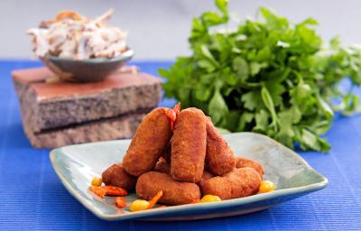 Foto da receita de croquete de carne, servidos em um prato acinzentado, sobre uma bancada azul com um maço de salsinha ao fundo