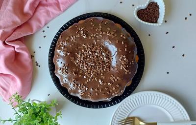 Foto em tons de marrom da receita do melhor bolo de cenoura servido sobre uma base preta com bastante cobertura de brigadeiro por cima com um paninho cor de rosa ao lado