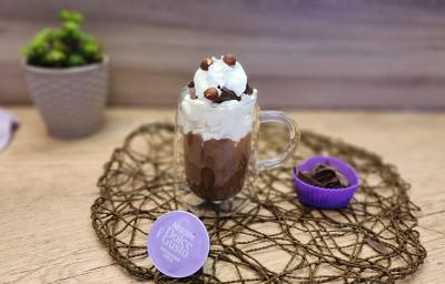 Fotografia em tons de lilás em uma bancada de madeira, com uma xícara alta de vidro e o milkshake de avelã com chocolate dentro dela. Em destaque, a cápsula de Mochaccino Avelã de Dolce Gusto e ao fundo um paninho lilás e um vasinho lilás com planta.