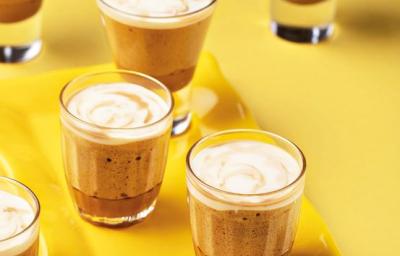 Fotografia em tons de amarelo em uma bancada de madeira amarela, um recipiente amarelo retangular e três copinhos de vidro com a mousse de café e caramelo dentro deles.