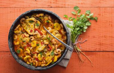 Fotografia em tons de vermelho em uma bancada de madeira vermelha com um recipiente redondo e fundo de pedra com a moqueca de peixe e camarão dentro dela.