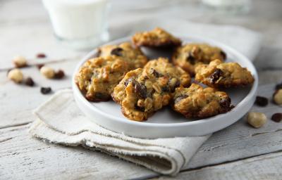 Fotografia em tons de branco, no centro, um prato branco com seis cookies com passas e grãos. Embaixo dos cookies, um pano de prato branco que está sobre uma mesa de madeira na cor branca.