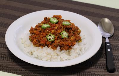 Fotografia em tons de marrom com um prato branco fundo ao centro. Dentro do prato existe uma porção de arroz coberta com carne moída e pedaços de quiabo. Ao lado existe uma colher.