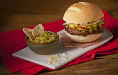 Fotografia em tons de vermelho em uma bancada de madeira com uma toalha vermelha, uma bandeja branca, um potinho com guacamole e nachos e um hambúrguer mexicano recheado com guacamole e nachos.