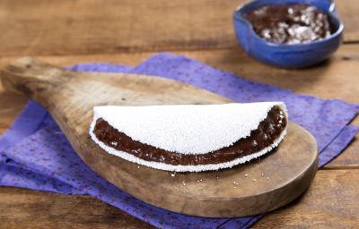 Fotografia em tons de marrom e roxo de uma bancada de madeira com um paninho roxo, sobre ele um tábua de madeira com uma tapioca. Ao fundo um recipiente roxo com creme de dark chocolate.