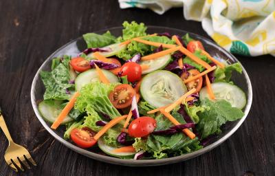 Fotografia de uma salada de folhas, pepino, cenoura ralada, tomates cereja e repolho roxo. Ao lado, um garfo na cor dourado, e ao fundo, uma tolha de mesa dobrada na cor branca, com desenhos em amarelo e verde, sobre uma mesa de madeira na cor preta.