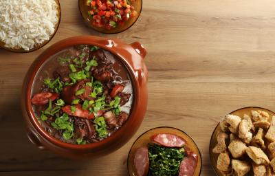 Fotografia vista de cima de uma feijoada completa, com cebolinha por cima, em um recipiente grande clássico de vidro marrom. Ao redor tem acompanhamentos de arroz, torresmo, vingrete e couve, todos em um recipiente de vidro, sobre uma mesa de madeira