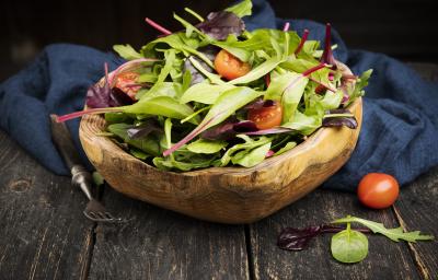 Fotografia em tons escuros de uma salada verde com salsão e tomates cereja dentro de um recipiente fundo de madeira, que está apoiado em um pano azul escuro, juntamente com um garfo de cabo preto, sobre uma mesa de madeira escura.