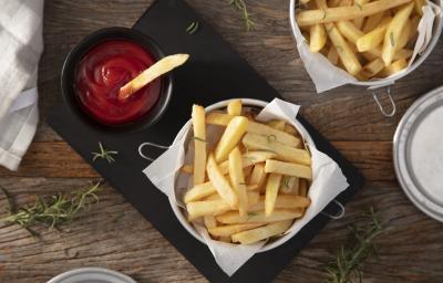 Fotografia em tons de marrom em uma bancada de madeira escura, uma tábua de ferro preta com um pote com batatas fritas dentro e um potinho ao lado com ketchup. Ao lado, um paninho e mais batata frita.