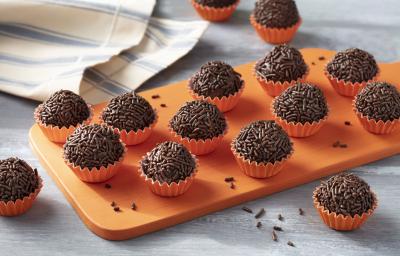 Imagem aproximada da receita de Brigadeiro Zero Lactose. Há diversos brigadeiros enrolados em forminhas em tom de laranja, todos sobre uma tábua de madeira em uma bancada decorada com um tecido em tom azul e bege