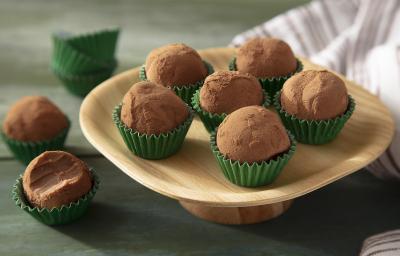 Fotografia em tons de marrom em uma bancada de madeira de cor verde. Ao centro, um prato de madeira contendo os brigadeiros. Ao, há um brigadeiro mordido e fundo, um pano branco.