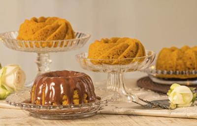 fotografia em tons de cinza, laranja e marrom vista de frente. Contém 4 bolos de cenoura e um deles esta com calda de chocolate, todos estão dentro de recipientes de vidros